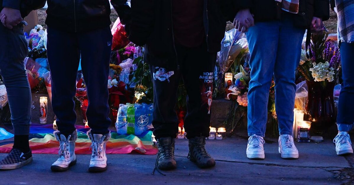 Photo with the legs of 5 people standing in front of an LGTBQ+ pride flag, flowers, and candles at dusk outside Club Q in Colorado Springs, CO after Nov 20, 2022 shooting.